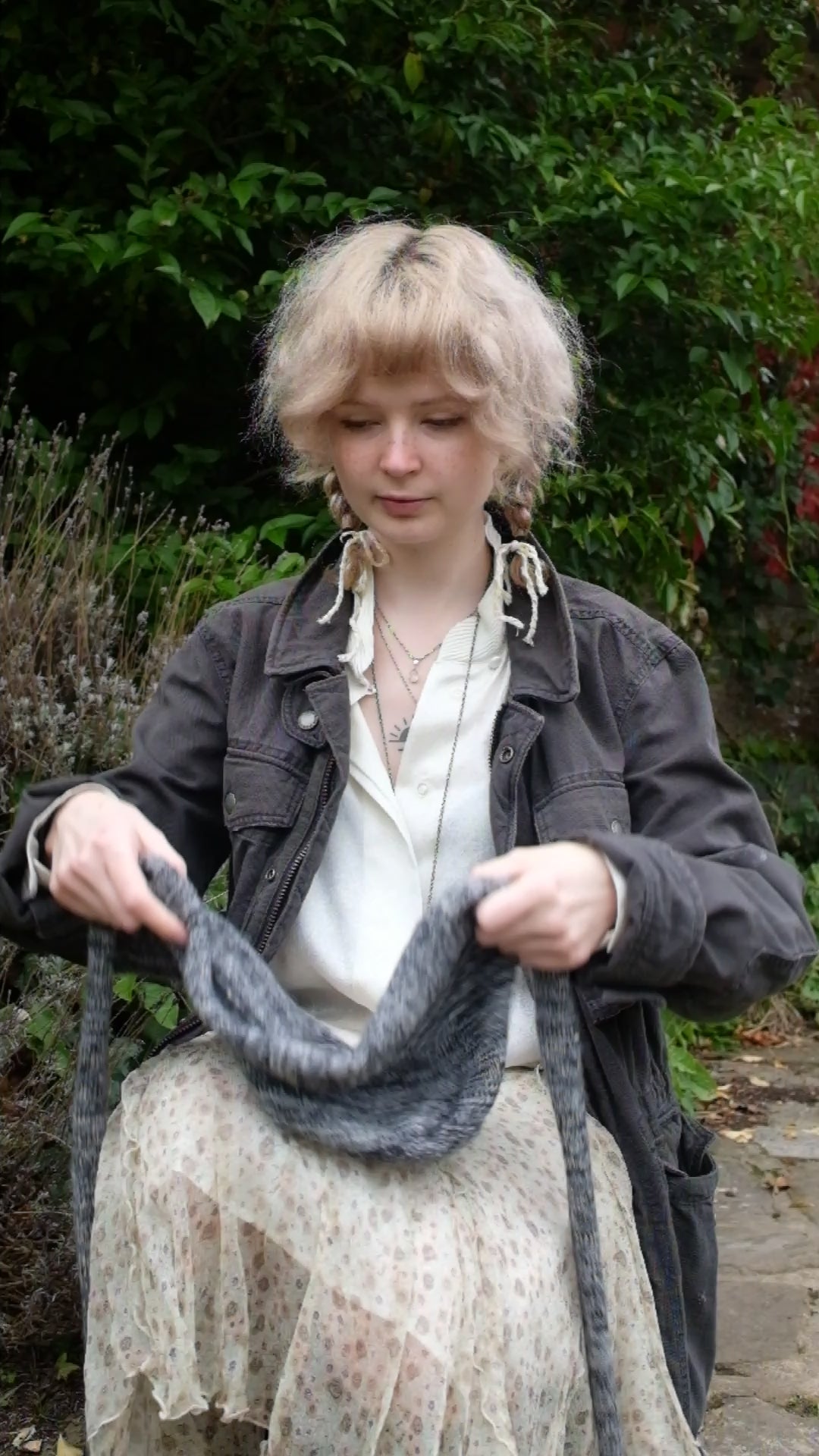 Person wearing a black mohair bonnet-style hood that fully covers the head and neck. With two long, soft extensions hanging down from the front. The hood is thick and textured, offering warmth and a cosy appearance, ideal for cold weather. The person is dressed in a light coloured coat and stands outdoors against a backdrop of green and red foliage 